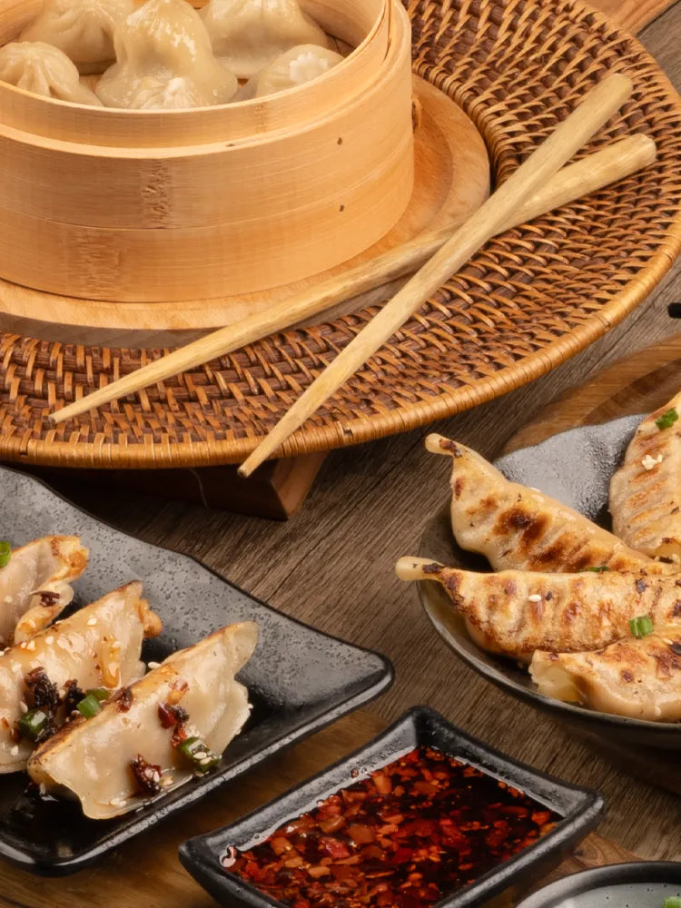 Close-up of pan-fried dumplings on black plates with chili dipping sauce, and steamed soup dumplings in a bamboo steamer, set on a wooden table with chopsticks.