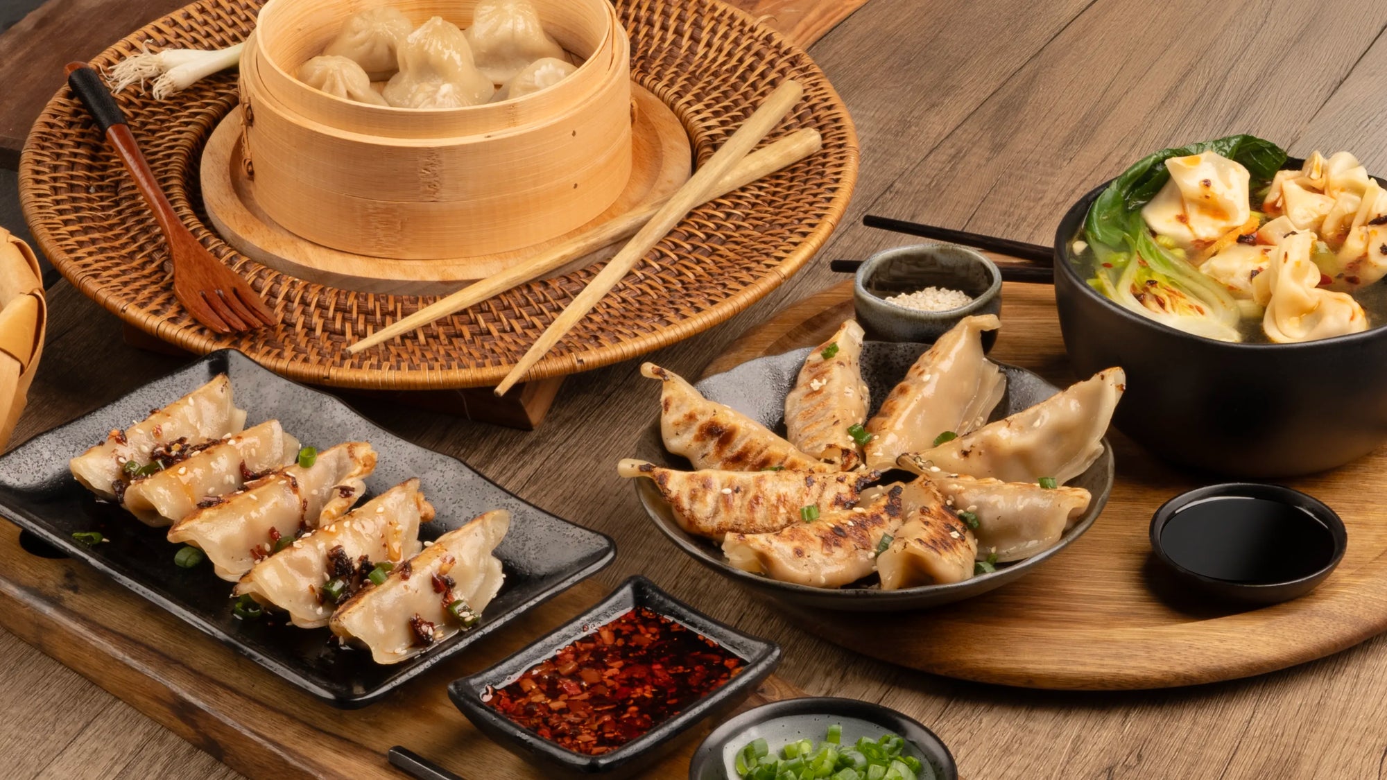 Assorted Chinese dishes on a wooden table, including soup dumplings in a bamboo steamer, pan-fried dumplings with dipping sauces, and a bowl of wonton soup with bok choy.
