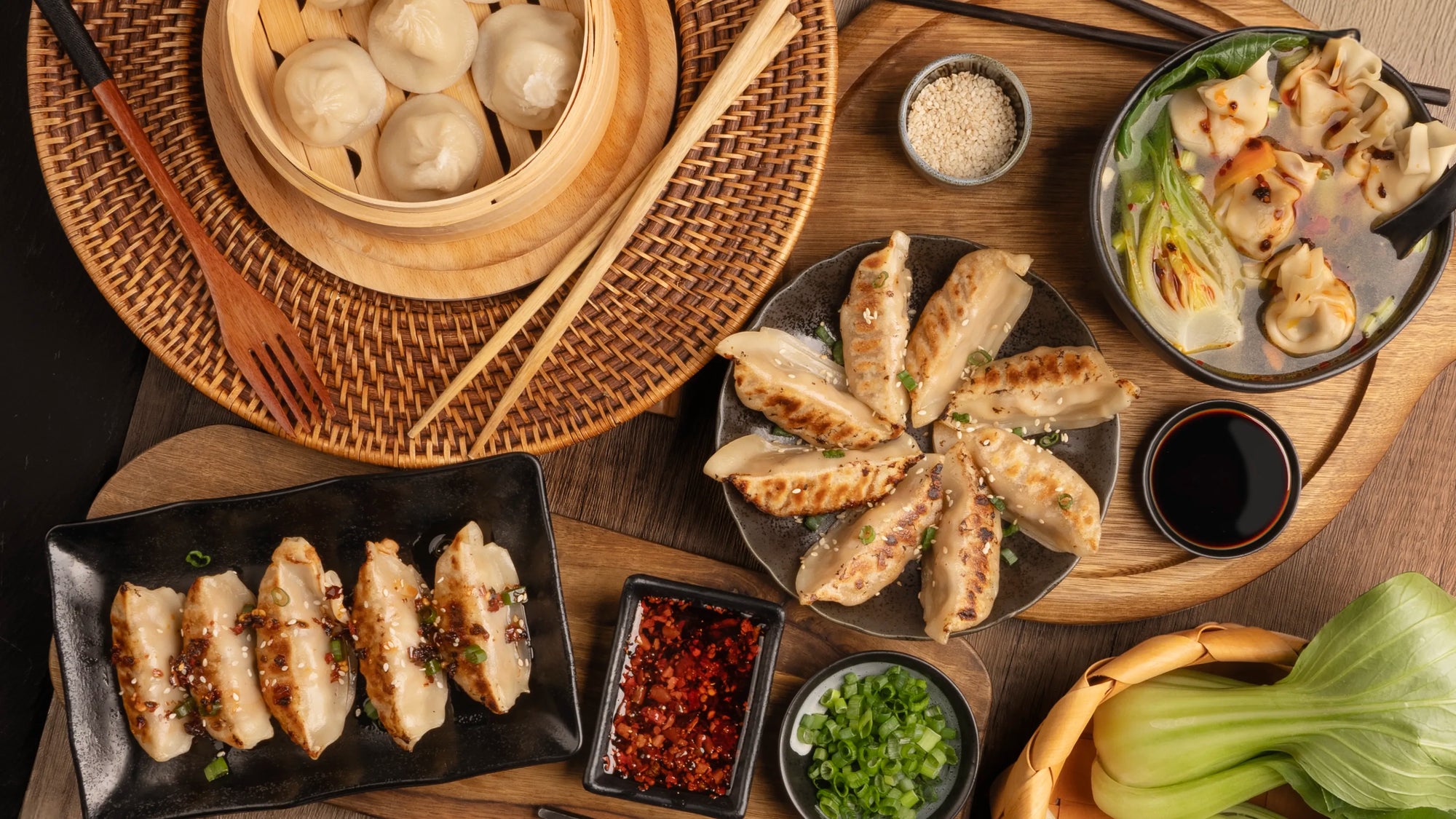 Top-down view of an Asian meal spread featuring pan-fried dumplings, steamed soup dumplings in a bamboo steamer, wonton soup, bok choy, dipping sauces, and garnishes on a wooden table.