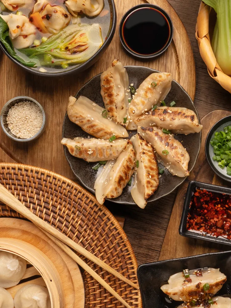 Top-down view of an Asian meal spread featuring pan-fried dumplings, steamed soup dumplings in a bamboo steamer, wonton soup, bok choy, dipping sauces, and garnishes on a wooden table.